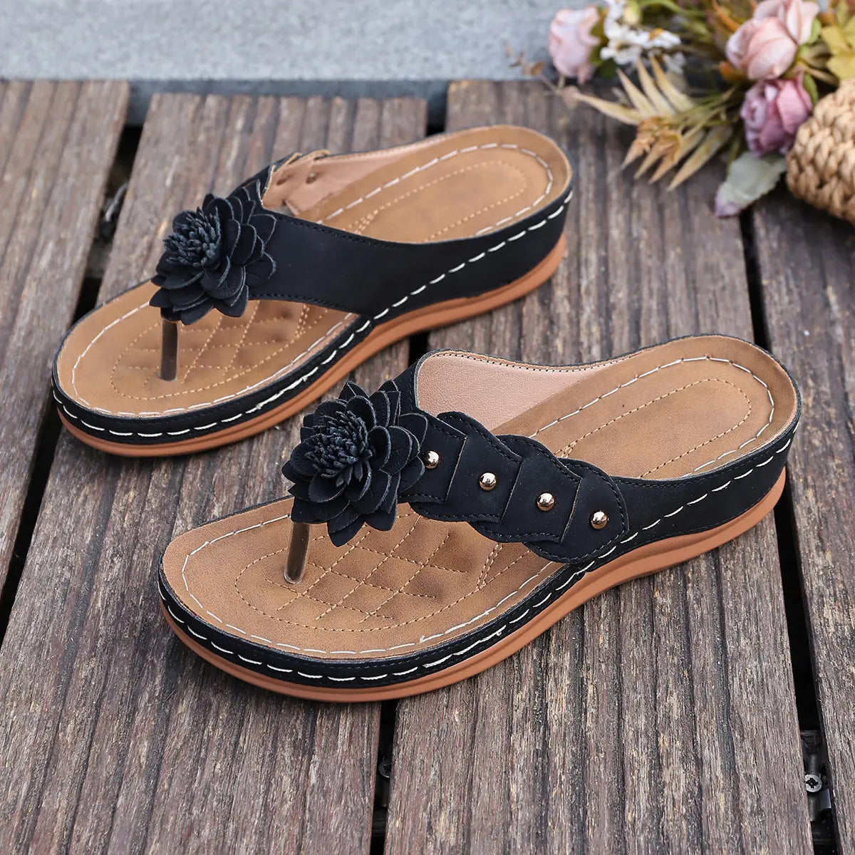 Black Lyraka thong sandals with layered fabric flower embellishments and gold studs, featuring white contrast stitching and a low wedge heel on a textured footbed. Shown on a rustic wooden surface.