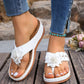 A pair of white thong sandals with fabric flower embellishments and gold studs, worn by a woman with blue jeans and a turquoise anklet, displayed on a wooden surface.