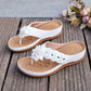 White Lyraka thong sandals adorned with layered fabric flowers and gold studs, displayed on a rustic wooden surface with a soft floral background.