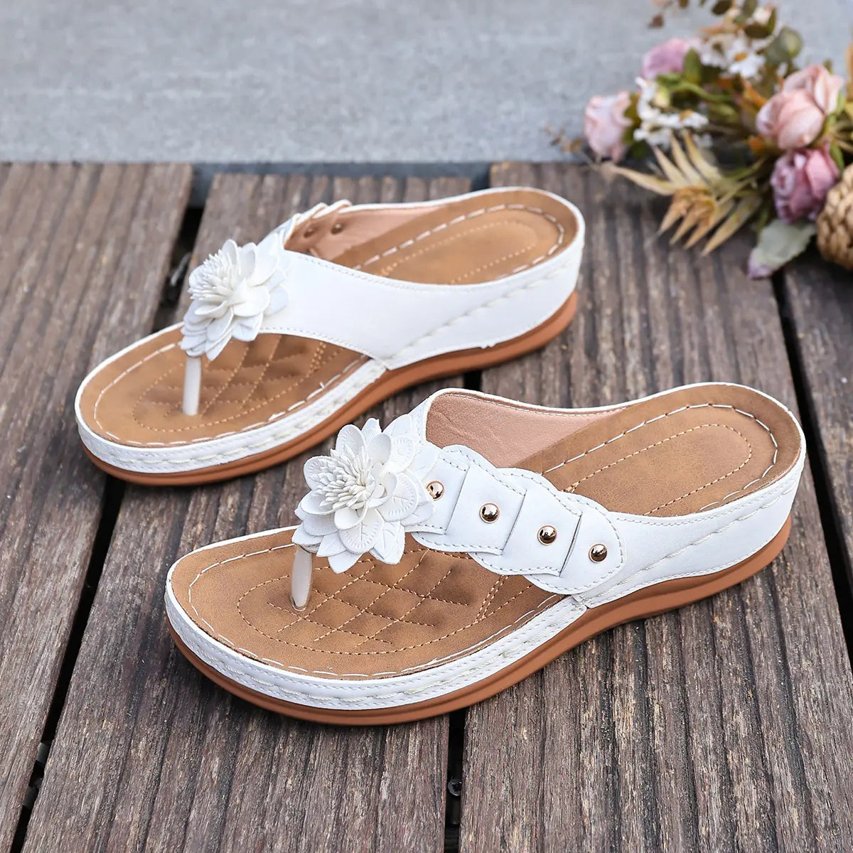 White Lyraka thong sandals adorned with layered fabric flowers and gold studs, displayed on a rustic wooden surface with a soft floral background.