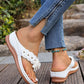 Woman's feet wearing white Lyraka thong sandals with layered fabric flower embellishments, gold studs, and white contrast stitching, paired with blue jeans and an anklet, on a wooden deck.
