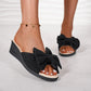 Black Lysa wedge slide sandals featuring a large decorative bow on the strap, shown on a model's feet with a gold anklet on a marble floor.