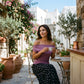 Woman in a dusty mauve off-shoulder top and black micro-floral maxi skirt, seated at an outdoor cafe table in a sunny European courtyard.