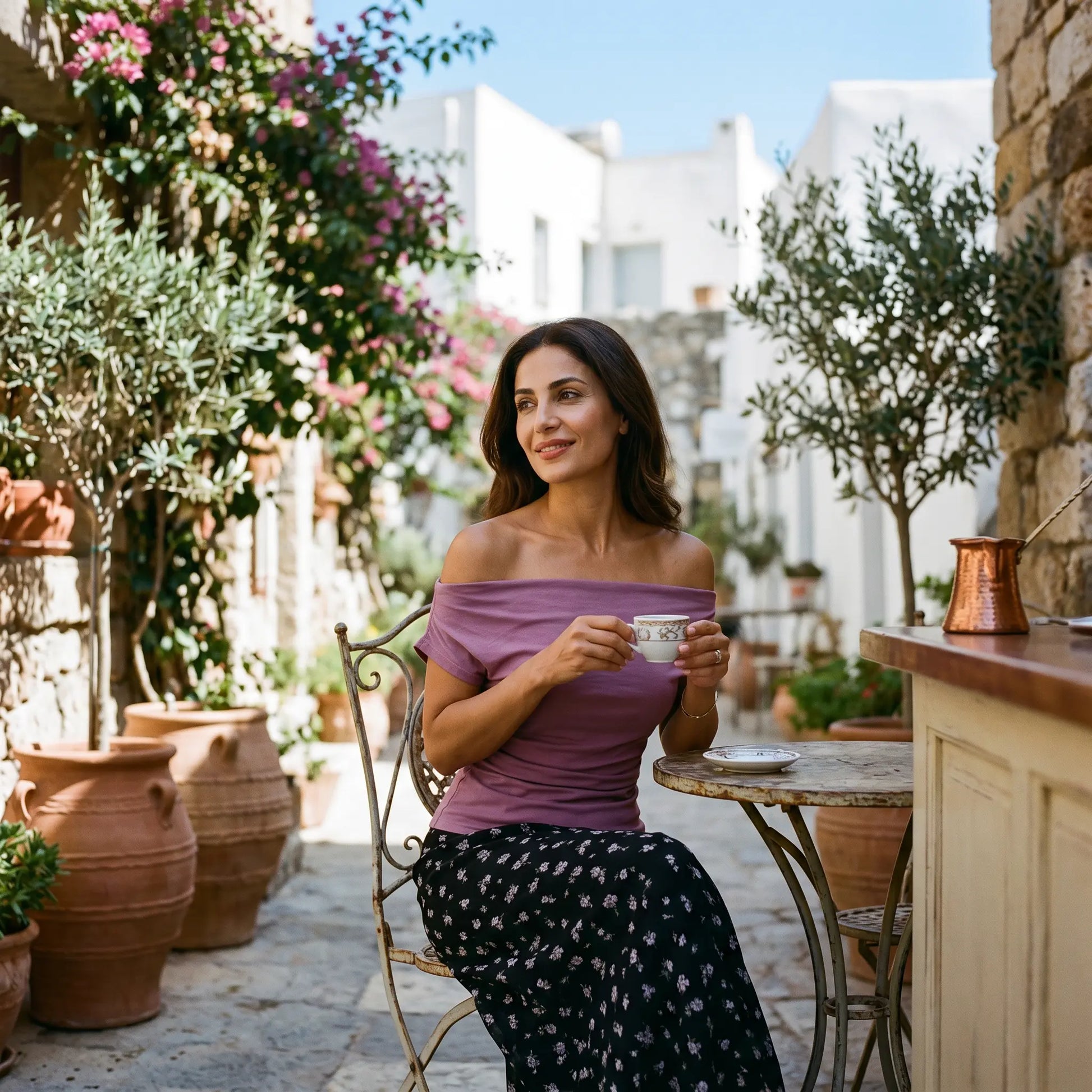 Woman in a dusty mauve off-shoulder top and black micro-floral maxi skirt, seated at an outdoor cafe table in a sunny European courtyard.