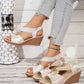 Shimmering gold metallic floral wedge sandals with elastic criss-cross straps and a decorative gold ring, shown on a model's foot with light blue jeans, alongside a second pair.