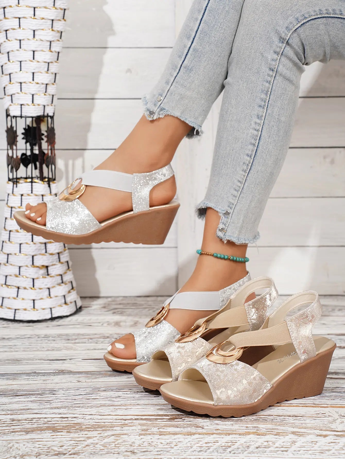 Shimmering gold metallic floral wedge sandals with elastic criss-cross straps and a decorative gold ring, shown on a model's foot with light blue jeans, alongside a second pair.
