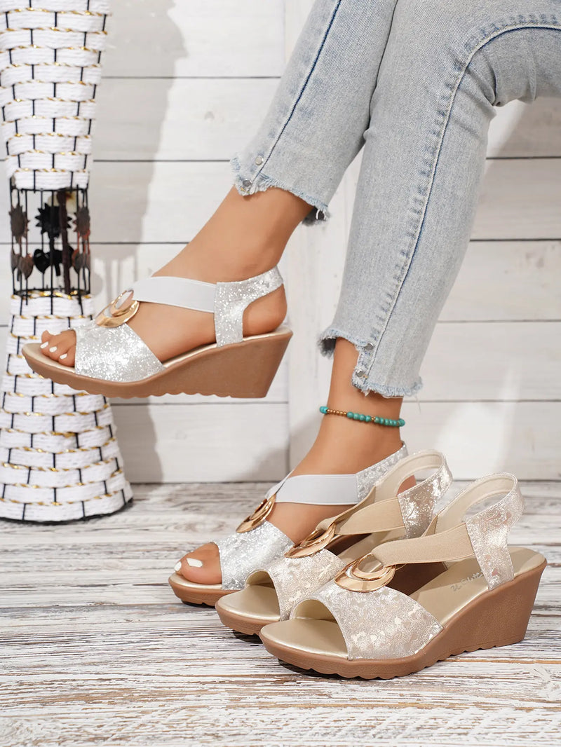 Shimmering gold metallic floral wedge sandals with elastic criss-cross straps and a decorative gold ring, shown on a model's foot with light blue jeans, alongside a second pair.