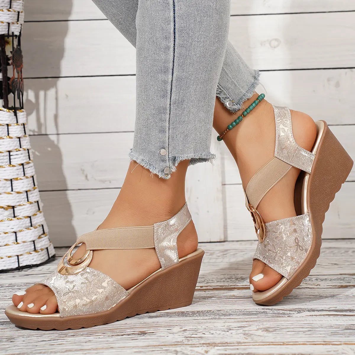 Elegant shimmering metallic floral wedge sandals with elastic criss-cross straps featuring a gold-tone ring, worn on a model's feet with distressed jeans against a white-washed wood plank background.