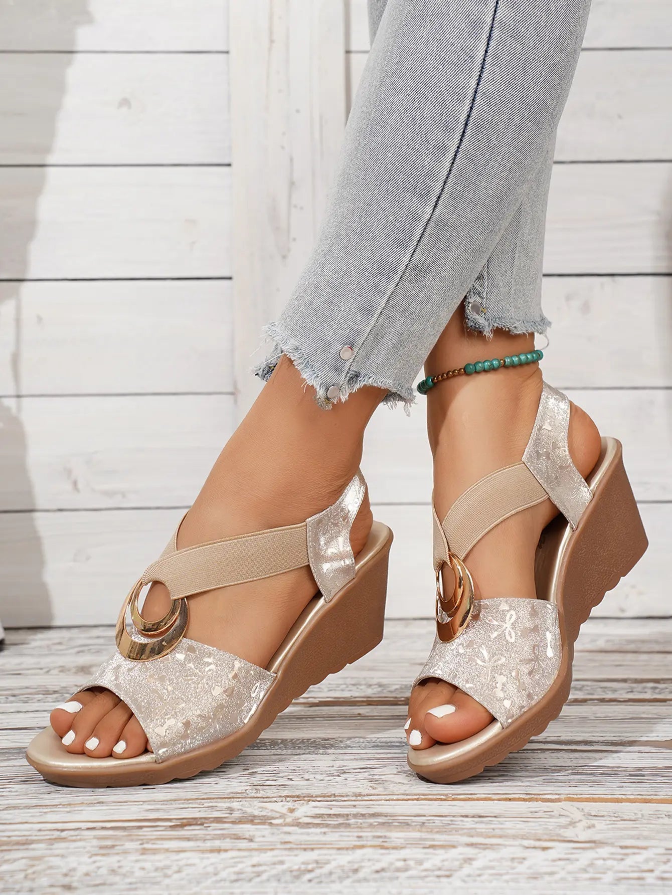 Shimmering metallic floral peep-toe wedge sandals with gold ring detail and elasticated straps, shown on a model's feet with light blue jeans.
