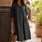 Woman wearing a Midnight Black V-neck midi dress with a crinkled texture, half sleeves, and front patch pockets, holding a straw bag against a decorative tiled wall in a courtyard setting.