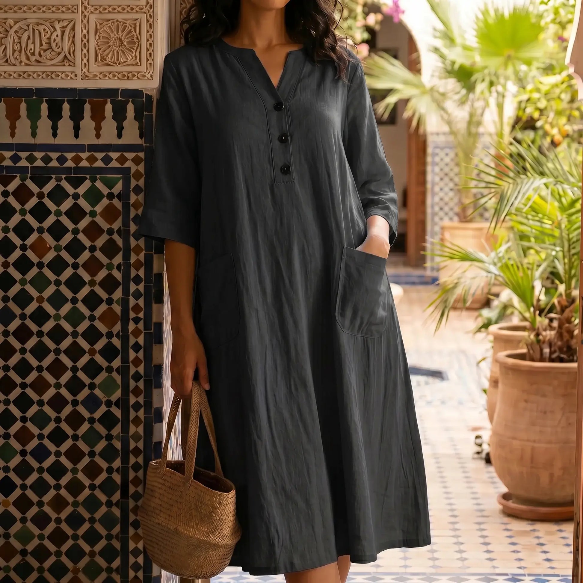 Woman wearing a Midnight Black V-neck midi dress with a crinkled texture, half sleeves, and front patch pockets, holding a straw bag against a decorative tiled wall in a courtyard setting.