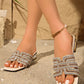 Close-up of beige Miravi embellished flat slide sandals adorned with geometric rhinestones, worn on a sandy surface with a hint of greenery.