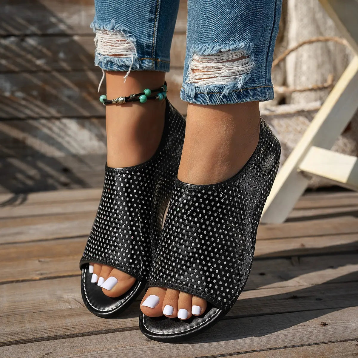 Miroka women's black perforated PU leather flat sandals with a peep-toe and single strap, worn with distressed jeans and a turquoise anklet on a wooden deck.