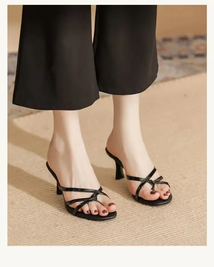 Elegant black strappy heeled mules with thin criss-cross straps, showcasing an open almond toe and slender stiletto heel worn by a model with black trousers.