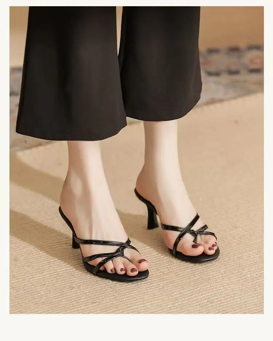 Elegant black strappy heeled mules with thin criss-cross straps, showcasing an open almond toe and slender stiletto heel worn by a model with black trousers.