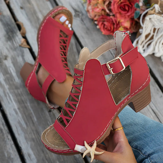 Red faux suede Roman sandals with a criss-cross lace-up front, peep-toe design, chunky block heel, and a buckle ankle strap, shown on a model's foot on a wooden deck.
