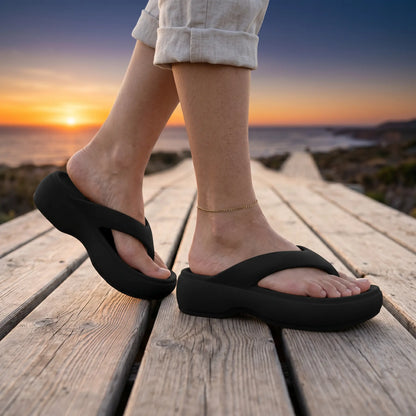 Woman walking on a wooden boardwalk at sunset, showcasing black Mivon elevated platform thong sandals made from matte EVA with a wide V-strap and 5cm thick sole, paired with a delicate gold anklet.