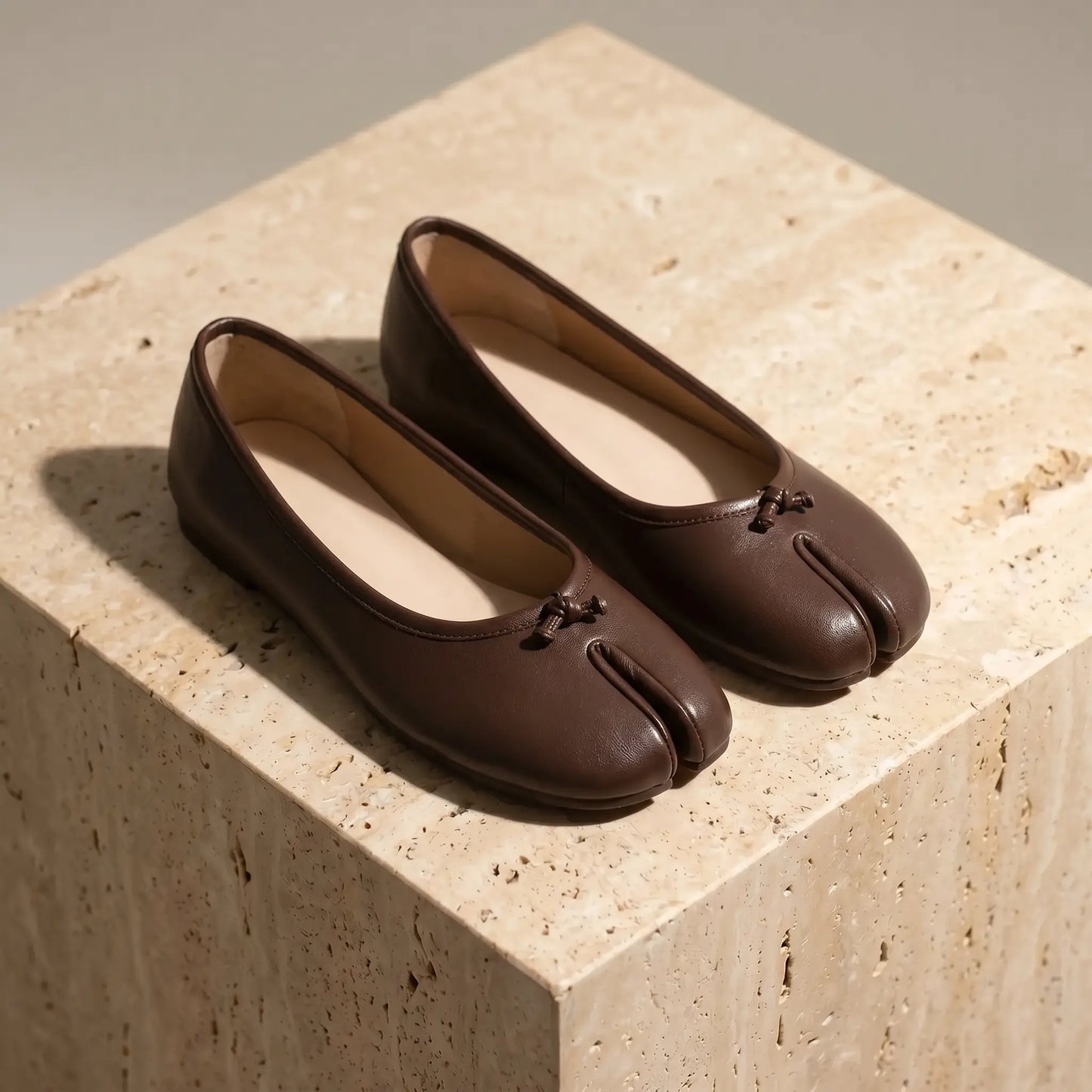 Lurran Mocha Brown vegan leather split-toe ballet flats, shown from a three-quarter view on textured stone. Features a delicate micro-bow at the toe cleft.