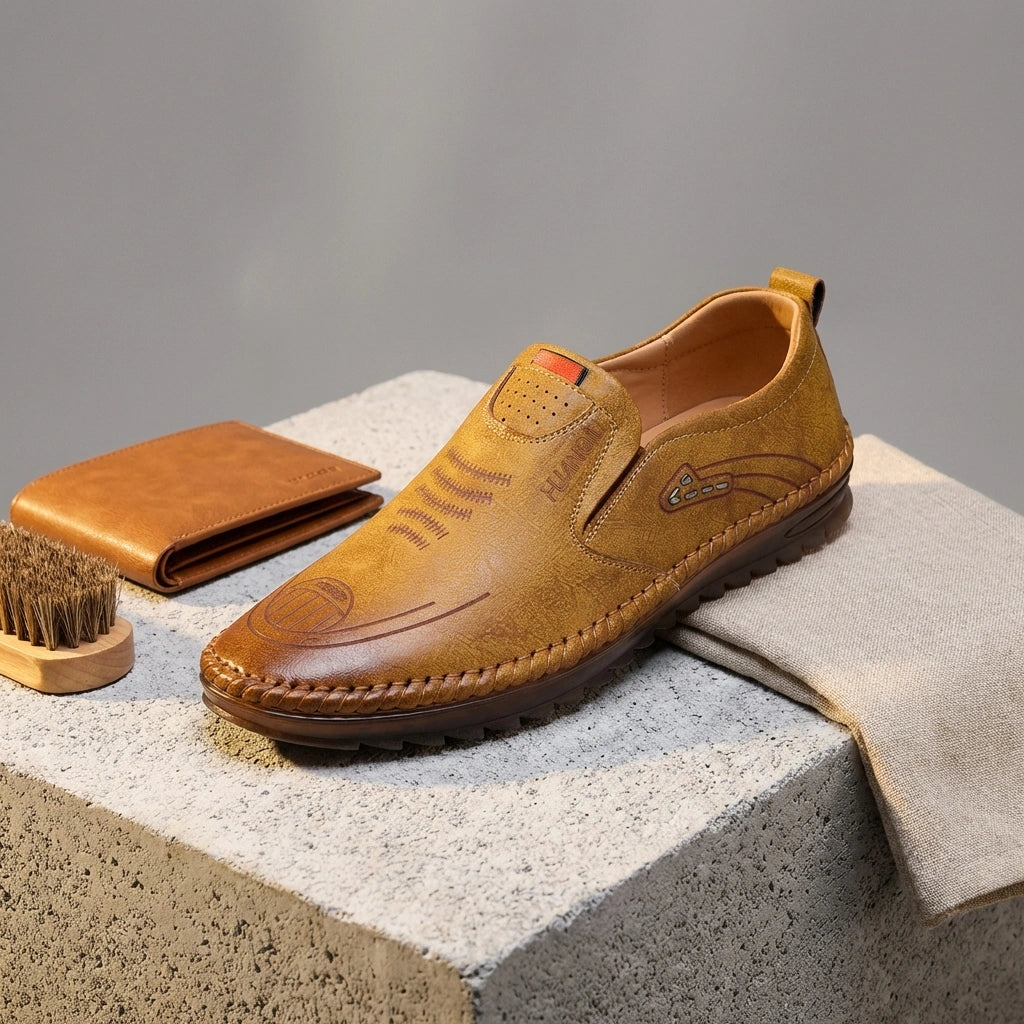 Mokasso men's comfort driving loafer in saddle brown vegan leather, styled on stone with wallet and brush — three-quarter view.