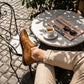 Mokasso men's hand-stitched driving loafer in saddle brown vegan leather worn casually at an outdoor cafe with espresso and newspaper.