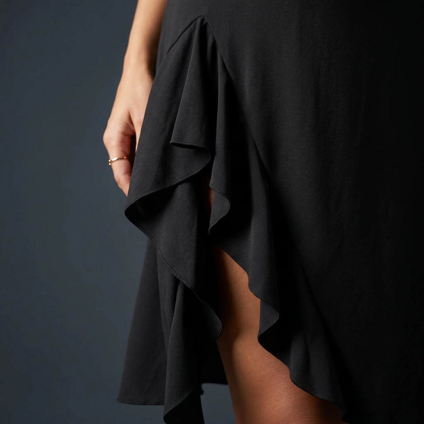 Close-up of the Mokazi black asymmetrical ruffle midi dress, highlighting the elegant cascading ruffles of the high-slit hem. Smooth matte jersey fabric drapes over a model's leg.