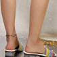 Model's feet wearing multi-color woven block heel slides with glitter heel and a gold anklet on a reflective surface.