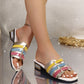 Model's feet wearing vibrant multi-color woven block heel slides with metallic gold, pink, blue, and yellow straps, styled with an anklet on a sequined background.