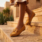 Mustard yellow faux-suede moccasin loafers with gold metallic bows, worn by a woman walking down sunlit stone steps. Highlights the flexible ridged rubber driving sole.