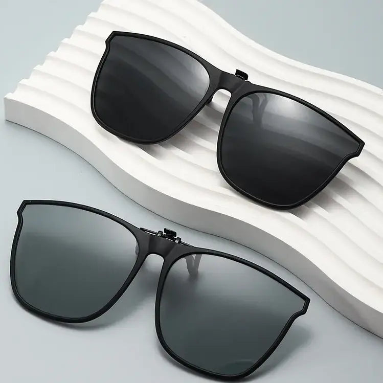 Two pairs of matte black metal clip-on sunglasses with square lenses, displayed from a front view on a textured studio surface.