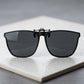 Matte black metal clip-on sunglasses with square polarized grey lenses, viewed from the front on a textured white surface.