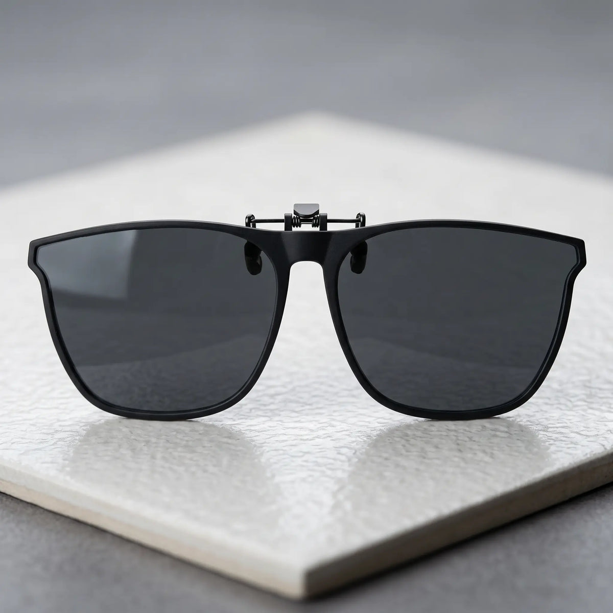 Matte black metal clip-on sunglasses with square polarized grey lenses, viewed from the front on a textured white surface.