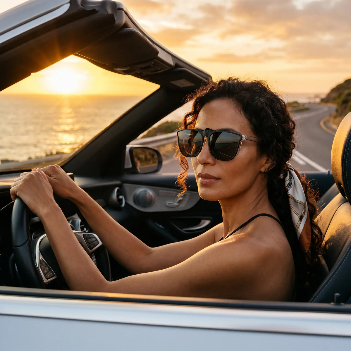 Woman wearing matte black square frame clip-on sunglasses while driving a convertible along a scenic coastal road at sunset.