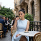Elegant woman in a Naliko powder blue sleeveless A-line mini dress, featuring a high round neckline and structured flared skirt, sips coffee at an outdoor cafe. This lifestyle image highlights the dress's timeless, romantic appeal.