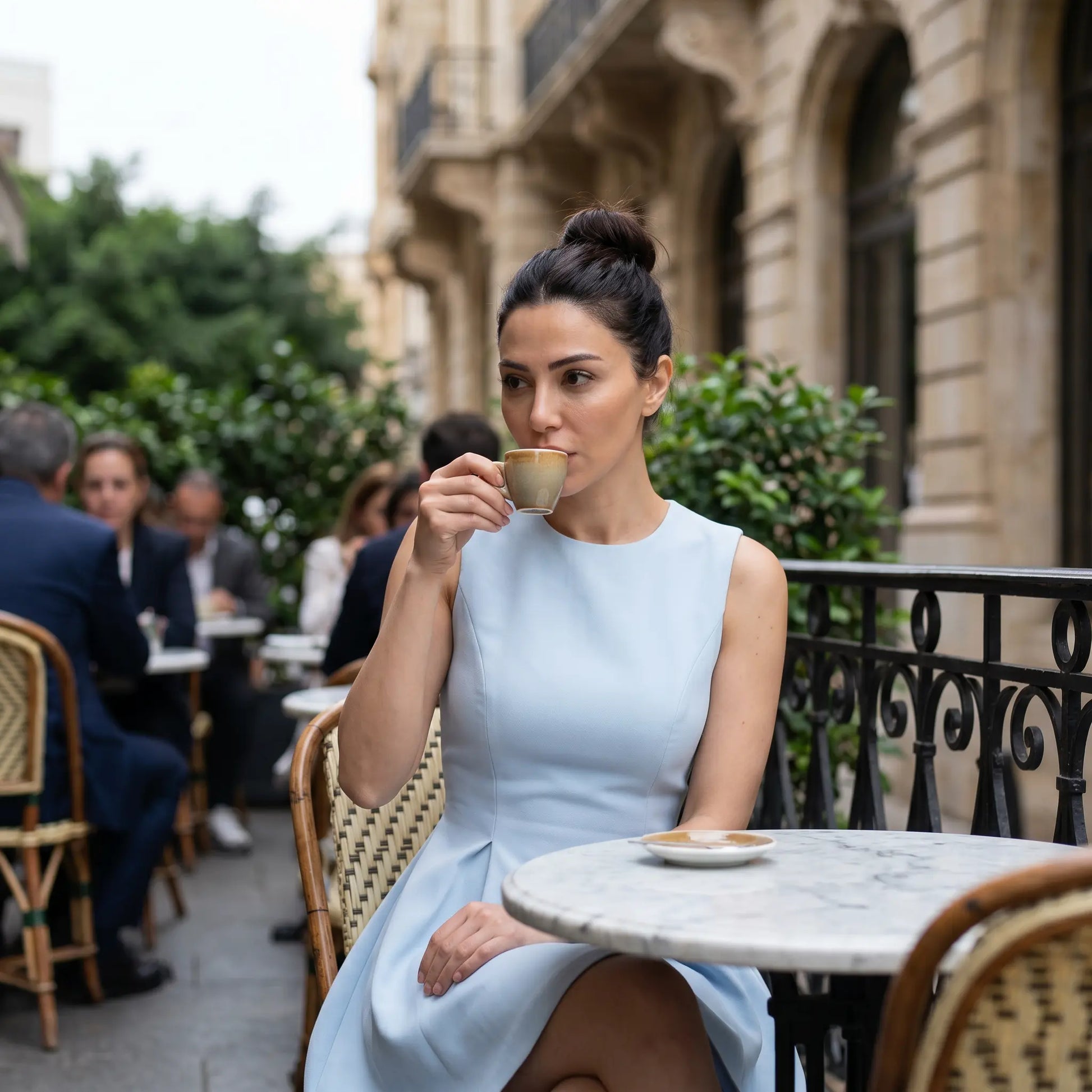 Elegant woman in a Naliko powder blue sleeveless A-line mini dress, featuring a high round neckline and structured flared skirt, sips coffee at an outdoor cafe. This lifestyle image highlights the dress's timeless, romantic appeal.