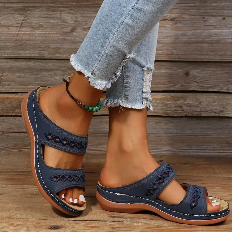 Navy blue women's slip-on slide sandals featuring double straps with geometric embroidered cut-outs and a subtle wedge heel, worn with distressed jeans.