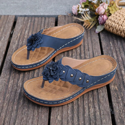 A pair of navy blue thong sandals adorned with a fabric flower embellishment and gold studs, resting on a wooden surface with blurred floral accents. These bohemian-style sandals feature white contrast stitching and a comfortable contoured footbed.