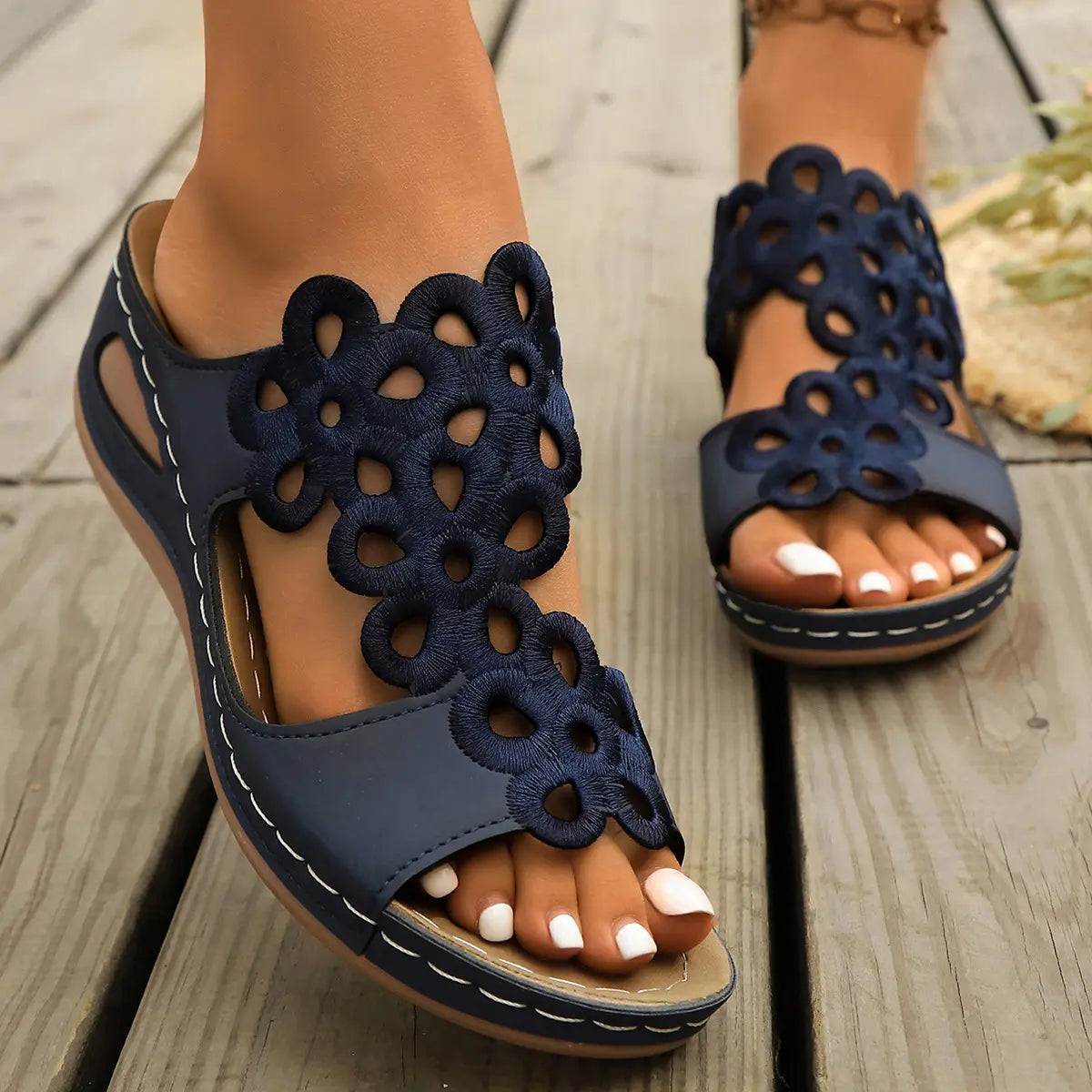 Women's navy blue bohemian wedge sandals featuring intricate laser-cut floral patterns on the straps, shown on feet with white-painted toenails against a rustic wooden background.