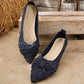 A pair of dark navy blue knit flat shoes with a subtle metallic thread sheen and a charming tied bow detail on the vamp, displayed from a three-quarter angle on a textured surface with rustic background elements.