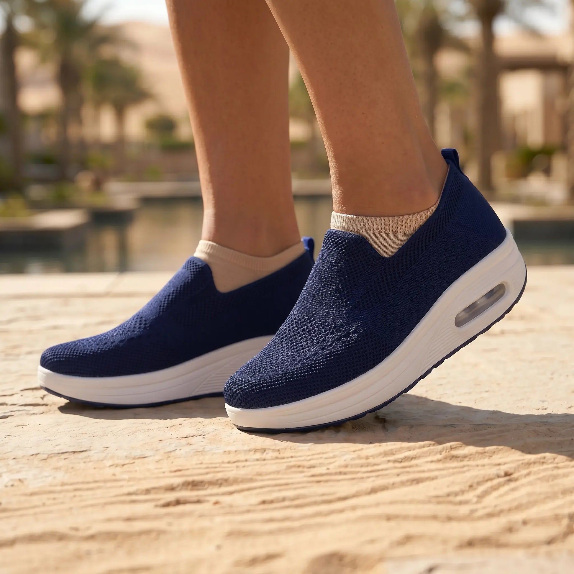 Exquisite navy blue knit slip-on sneakers, featuring a thick white platform sole with a visible heel air cushion, worn outdoors on a sandy path. The breathable upper provides effortless comfort.