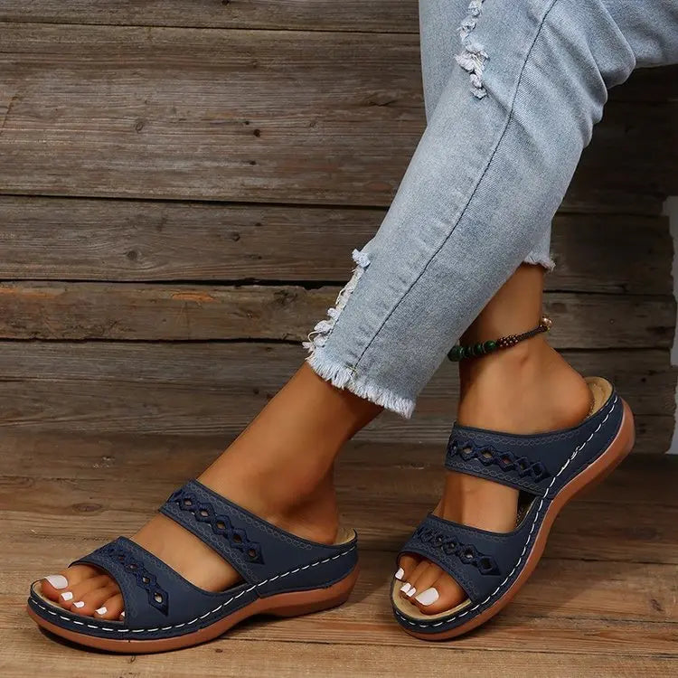 Close-up of a woman's feet wearing navy blue slip-on slide sandals with intricate geometric embroidery on the straps and a contoured wedge sole, paired with distressed denim jeans.