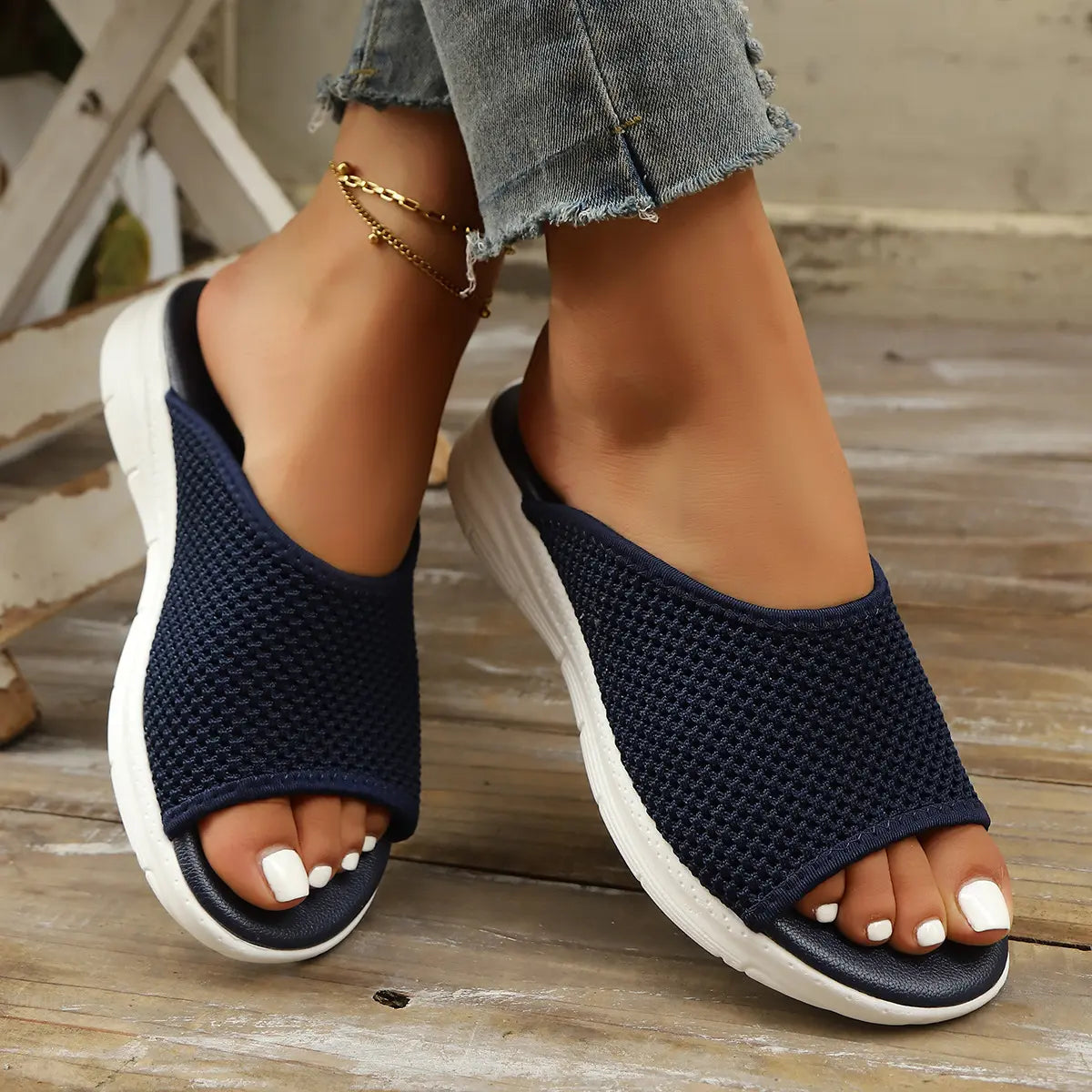 Close-up of a woman's feet wearing navy blue Kithra breathable knit slide sandals with white-painted toenails and a delicate gold anklet.