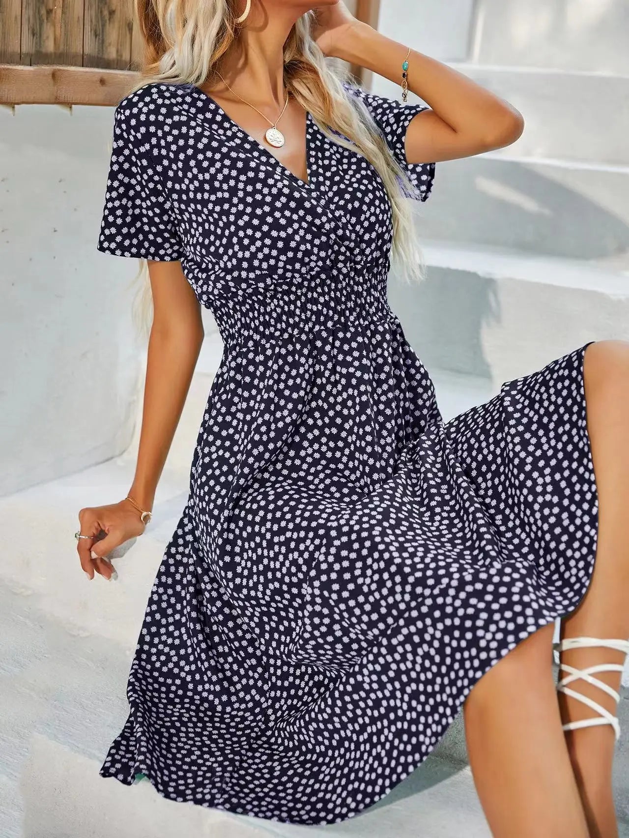 A model wears a navy ditsy floral V-neck midi dress with short flutter sleeves and a smocked cinched waist, styled with a necklace and bracelet.