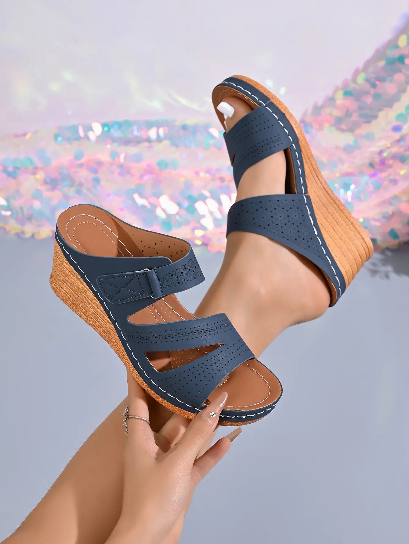 Navy blue perforated leather wedge sandals with adjustable hook-and-loop straps and white contrast stitching, shown on a model's foot with a bokeh background.