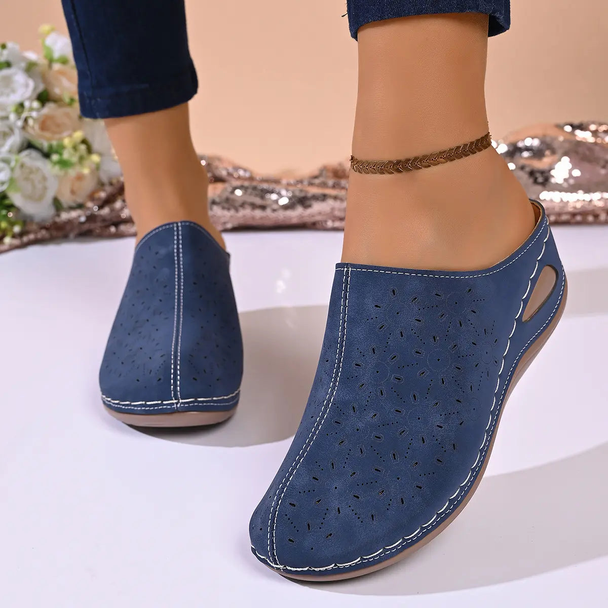 Close-up of women's navy blue perforated wedge mules worn on feet, showcasing intricate floral cut-outs and white stitching details on the slip-on design.