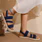 Navy blue elasticated woven strap flat sandals worn by a model with a flowing white dress, showcasing the fish-mouth toe and braided sole trim. These comfortable summer sandals feature a rear ankle strap and are styled in a bohemian lifestyle setting.