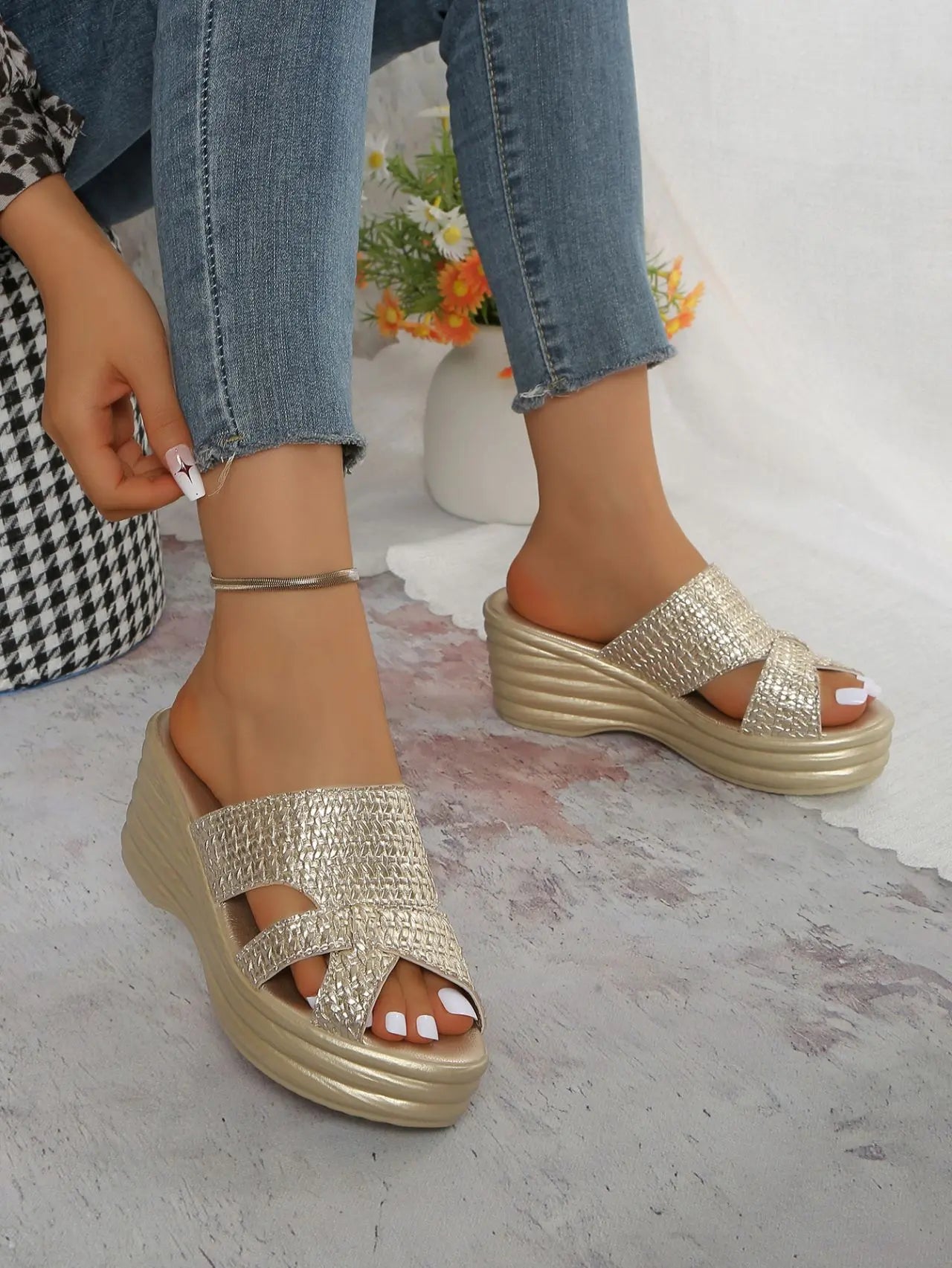 Shimmering gold platform wedge sandals worn by a model, featuring a woven texture, peep-toe design, and a high wedge heel. The footwear is styled with blue denim jeans and an anklet.