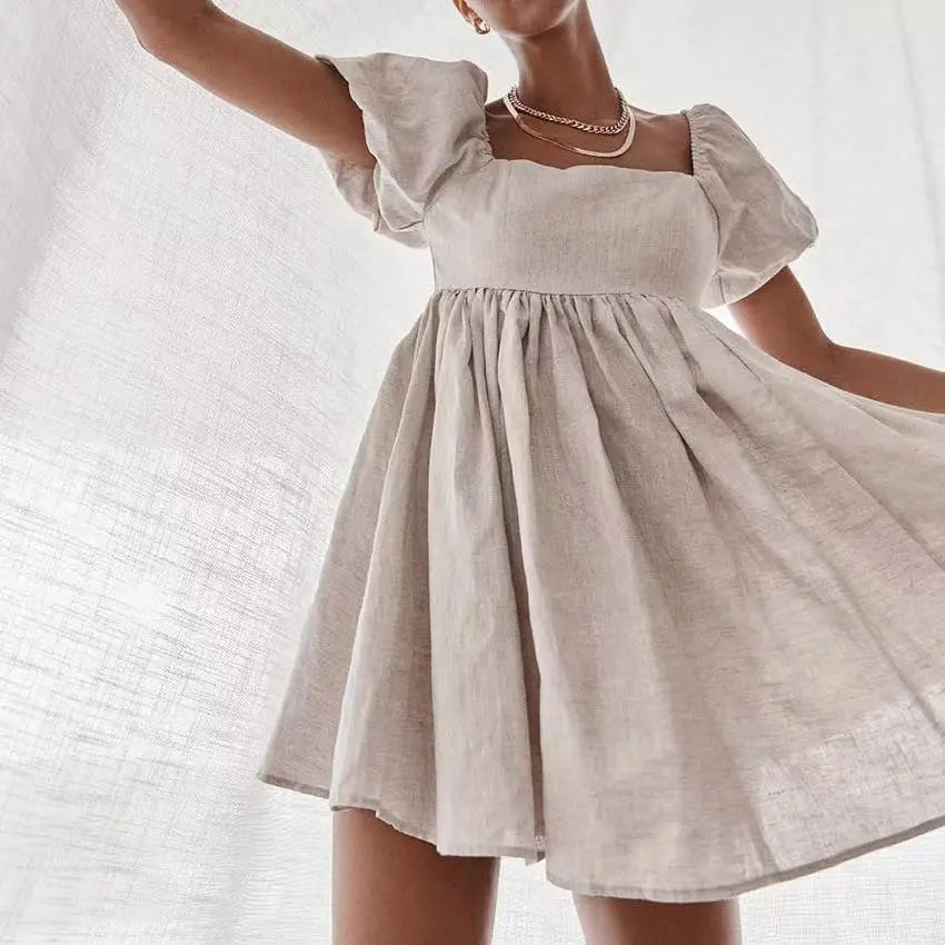 A woman models a short, A-line beige linen-blend dress with a square neckline and voluminous puff sleeves, shown in a flowing pose against a textured light background.