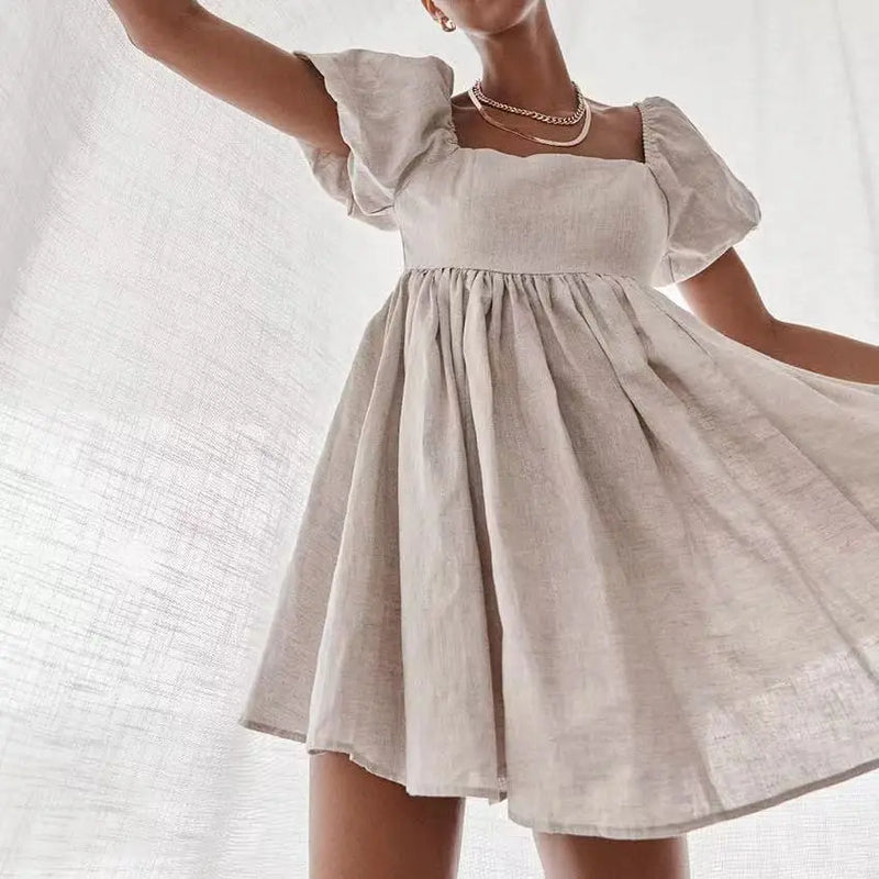 A woman models a short, A-line beige linen-blend dress with a square neckline and voluminous puff sleeves, shown in a flowing pose against a textured light background.
