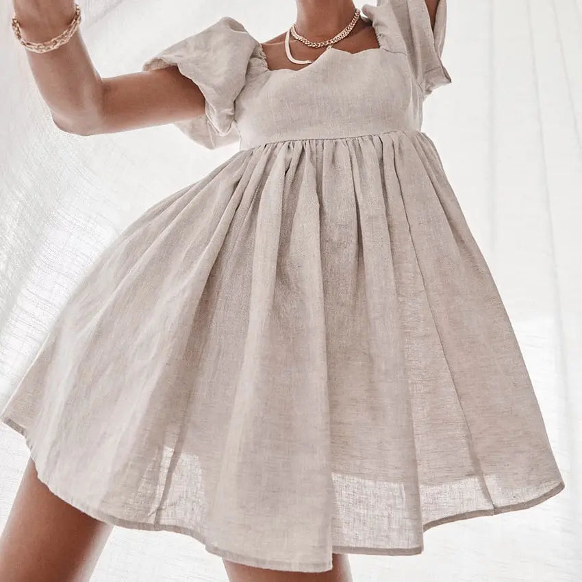 Nimae short beige linen blend dress with square neckline, voluminous puff sleeves, and gathered A-line mini skirt, worn by a model against a soft white curtain.