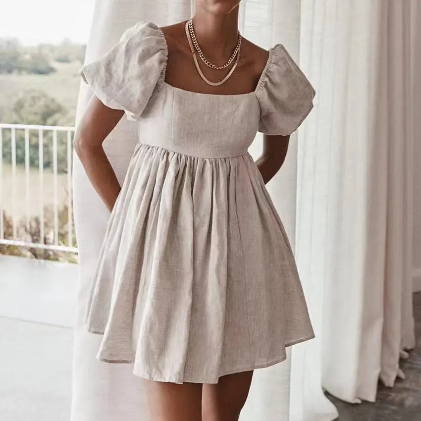 A woman wearing the Nimae square neck puff sleeve mini dress in warm beige linen blend, showcasing its voluminous sleeves, gathered waist, and A-line skirt. The model stands near a window with soft light.